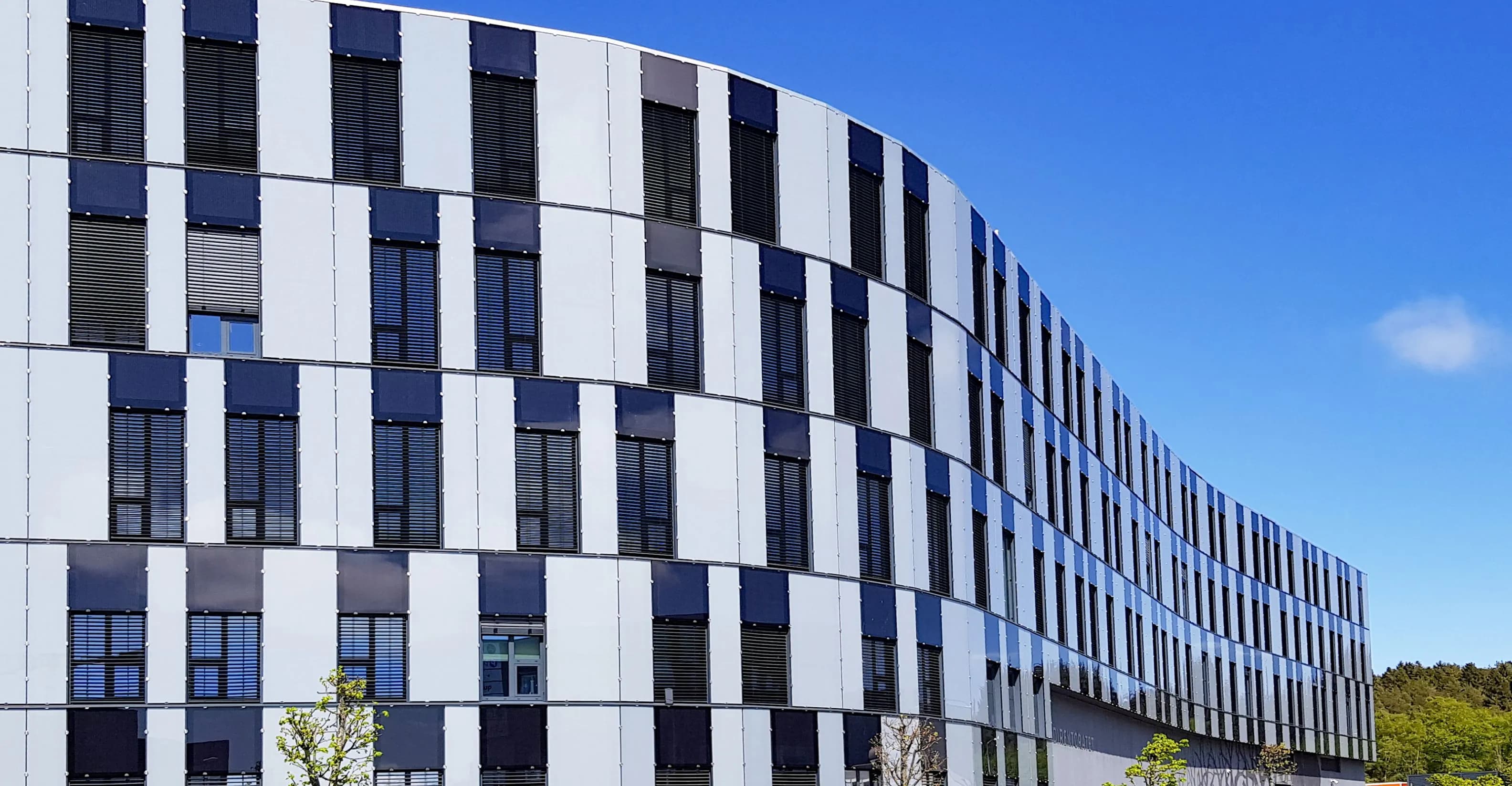 Wide view of a modern dark-gray office building with grid panels and rows of bronze-framed windows, pale-green vertical accents, and a lamppost, set against a clear blue sky.