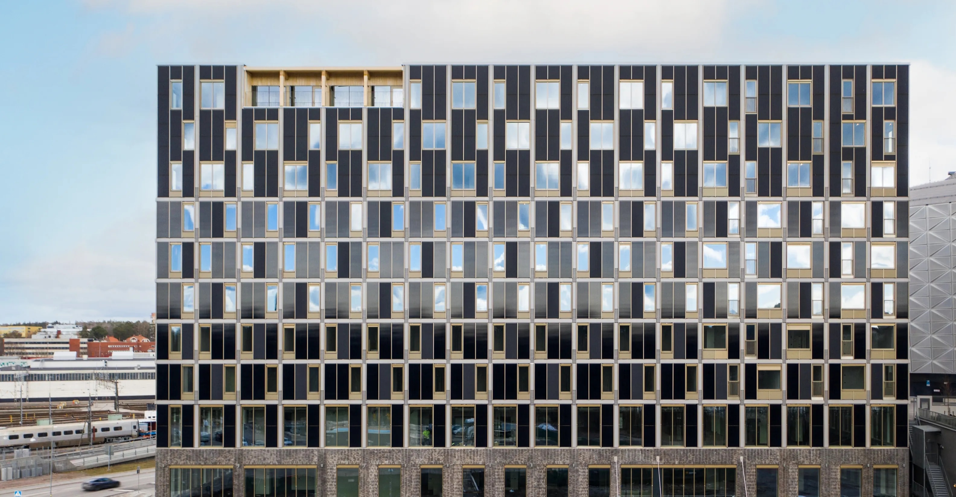 Contemporary hotel building with a geometric black and gold facade featuring large reflective windows, located near railway tracks and modern urban infrastructure.