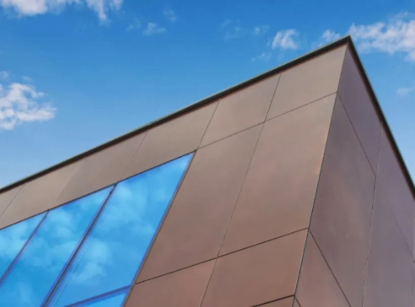 Close-up of a building corner clad in copper-brown facade panels with tall blue reflective windows against a clear blue sky.